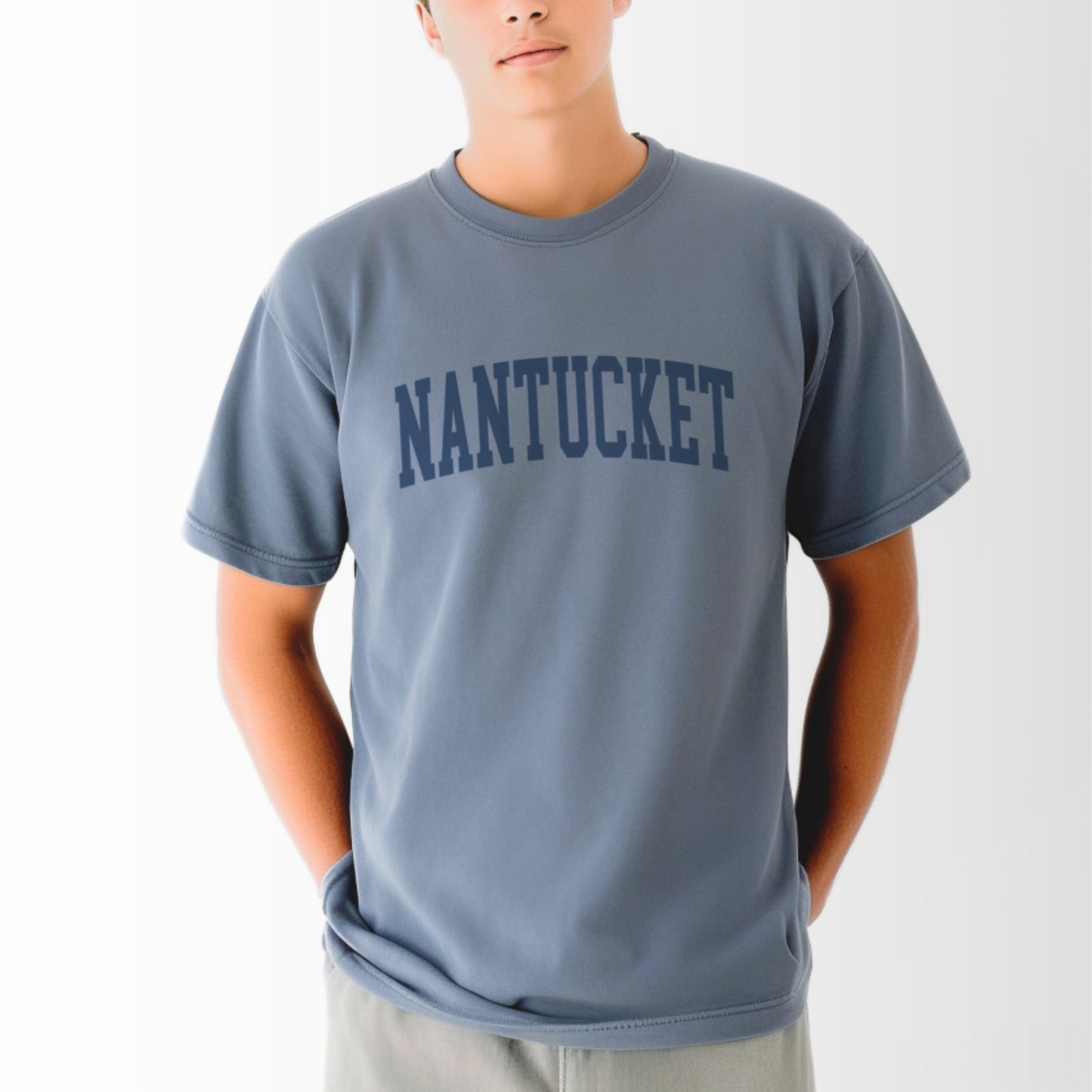 A model wears the Blue Jean Adult Unisex Nantucket Massachusetts Varsity Comfort Colors T-shirt.  The ﻿Nantucket Massachusetts Varsity﻿ graphic is in bold Navy in a Collegiate Block style.