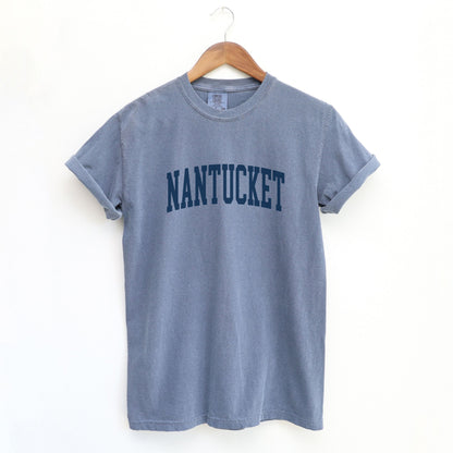 The Blue Jean Adult Unisex Nantucket Massachusetts Varsity Comfort Colors T-shirt lays flat on a white background. The ﻿Nantucket Massachusetts Varsity﻿ graphic is in bold Navy in a Collegiate Block style.