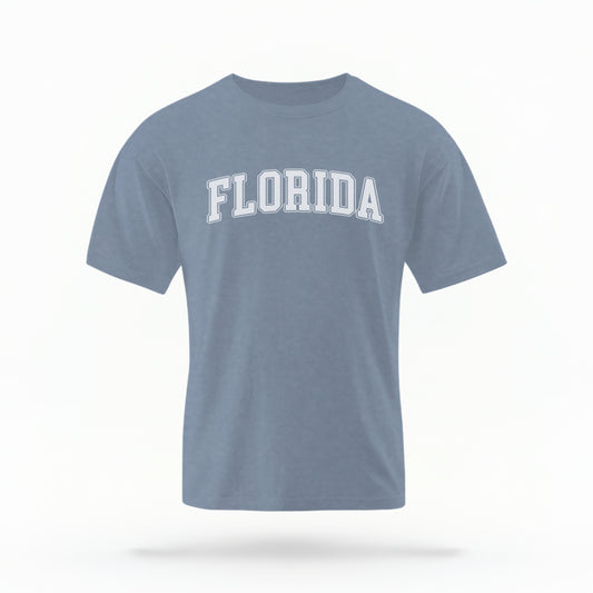The blue jeans Adult Unisex Florida Varsity Comfort Colors T-shirt lays flat on a white background. The Florida graphic is in bold white in a varsity offset style.