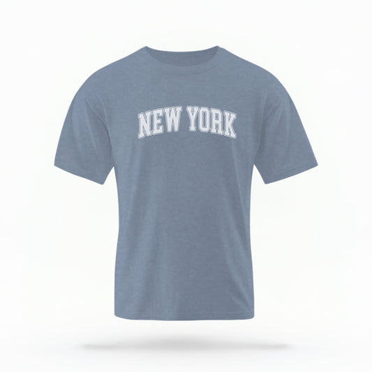 The Blue Jeans Unisex New York Varsity Comfort Colors T-shirt lays flat on a white background. The New York graphic is in bold white in a varsity offset style.