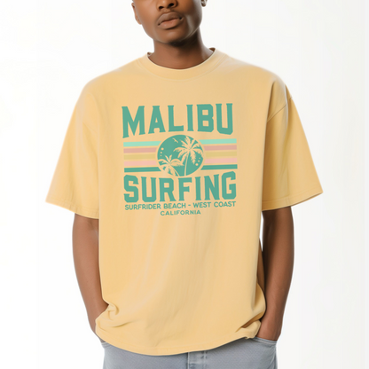 A model wears the Butter Adult Unisex Malibu California Surfing Comfort Colors T-shirt.  The ﻿Malibu California Surfing﻿ graphic is in bold Lifestyle in a Multicolor style.