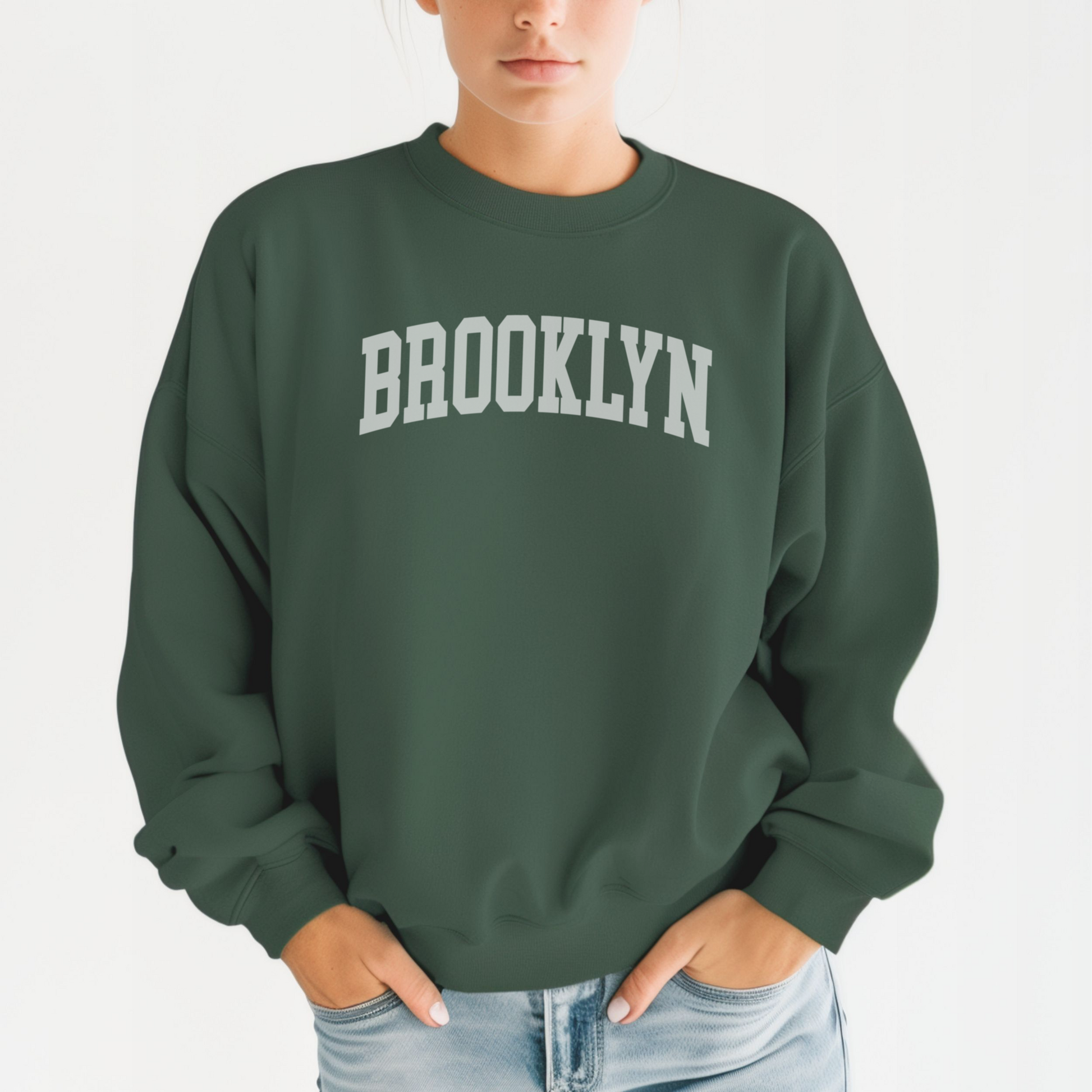 A model wears the Forest Green Adult Unisex Brooklyn NY Collegiate Crewneck Sweatshirt.  The ﻿Brooklyn NY Collegiate﻿ graphic is in bold White in a Varsity style.