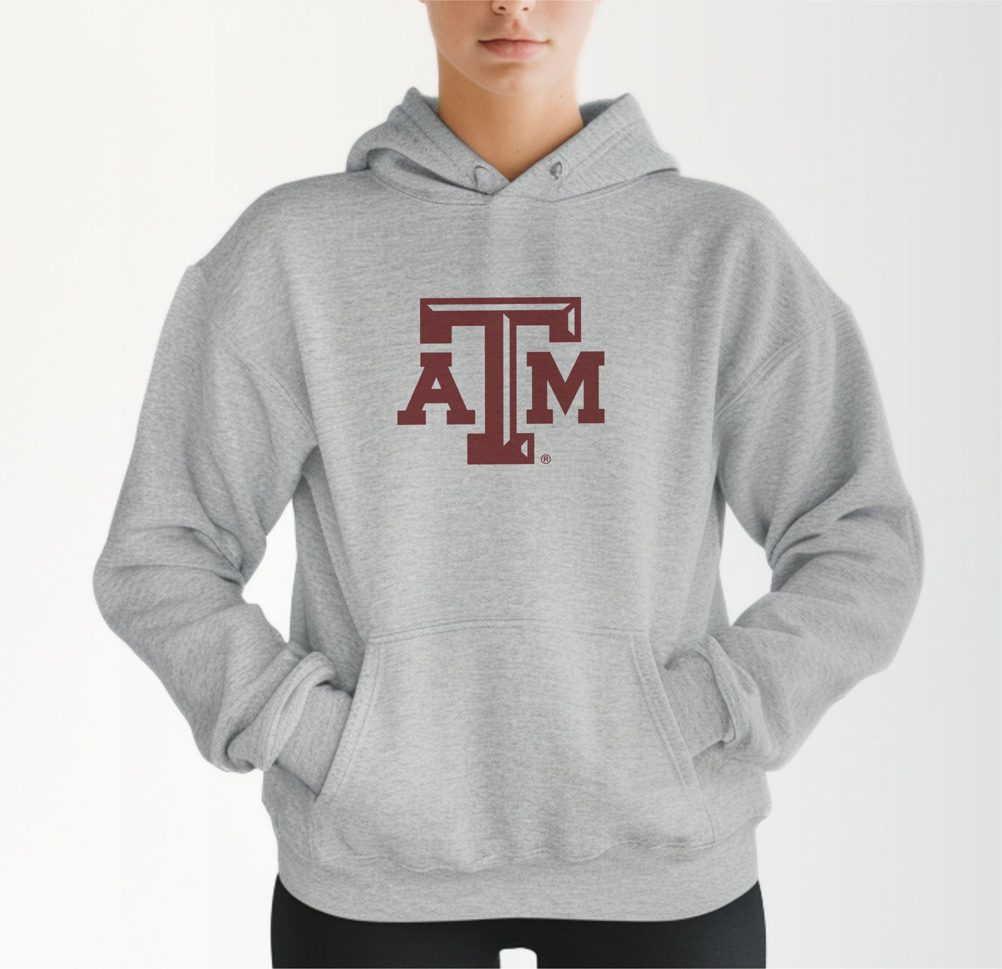 A model wears the Sport Grey Adult Unisex Texas A&M Logo Hooded Sweatshirt.  The ﻿Texas A&M Logo﻿ graphic is in bold Maroon in a Varsity style.