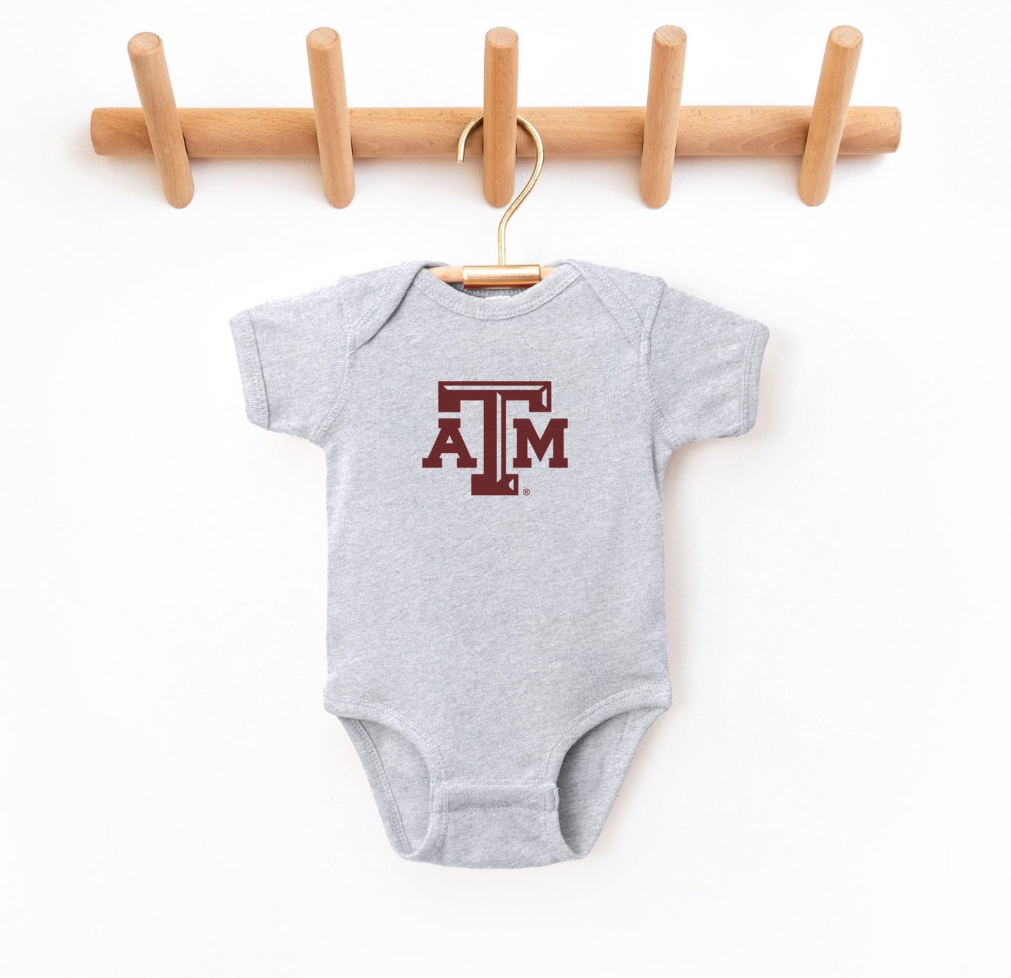 The Heather Grey Infant Unisex Texas A&M Logo Bodysuit lays flat on a white background. The ﻿Texas A&M Logo﻿ graphic is in bold Maroon in a Varsity style.