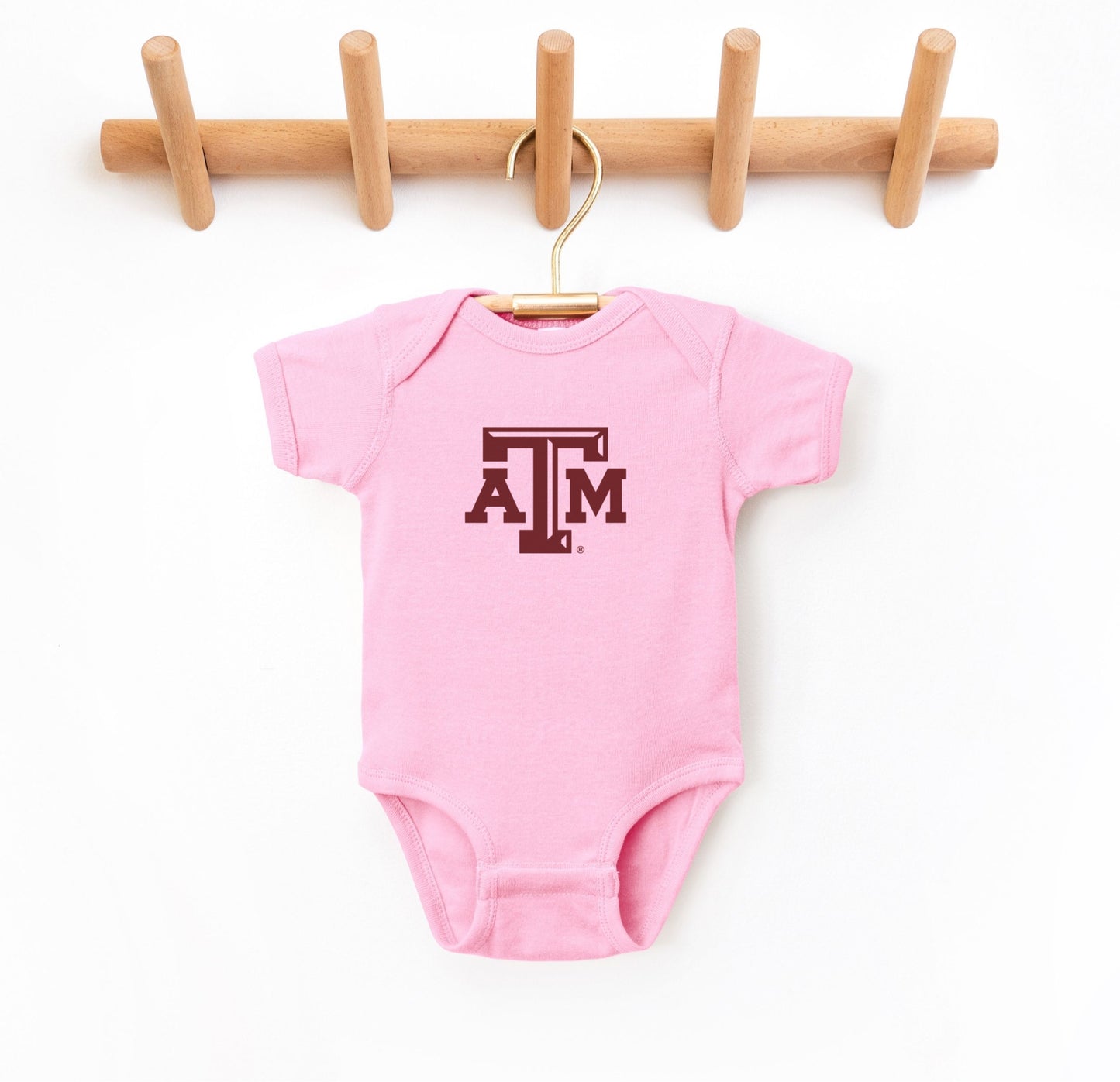 The Pink Infant Unisex Texas A&M Logo Bodysuit lays flat on a white background. The ﻿Texas A&M Logo﻿ graphic is in bold Maroon in a Varsity style.