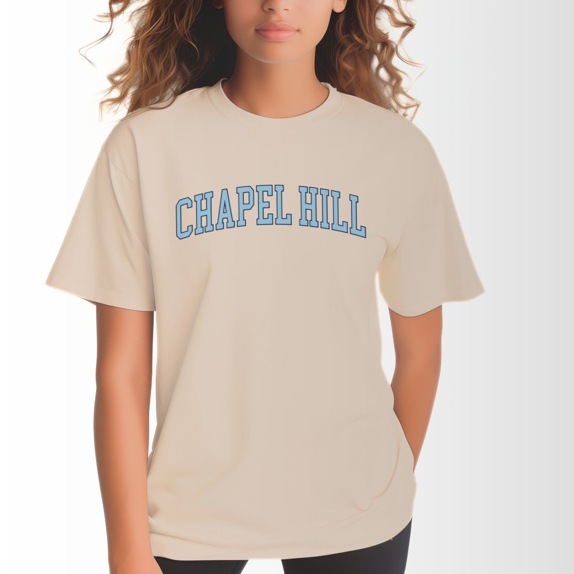 A model wears the Ivory Adult Unisex Chapel Hill Varsity Comfort Colors T-shirt.  The ﻿Chapel Hill Varsity﻿ graphic is in bold Lifestyle in a Collegiate style.