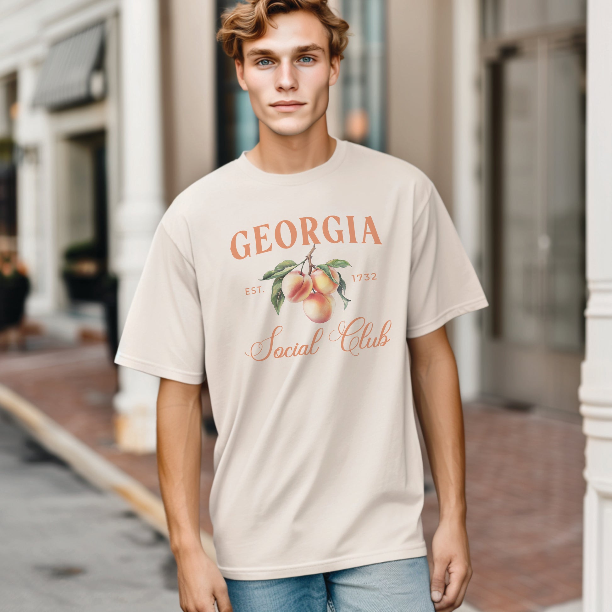 A model wears the Ivory Adult Unisex Georgia Peach Social Club T-shirt.  The Georgia Peach  graphic is in bold orange and green in a  preppy old money style. 