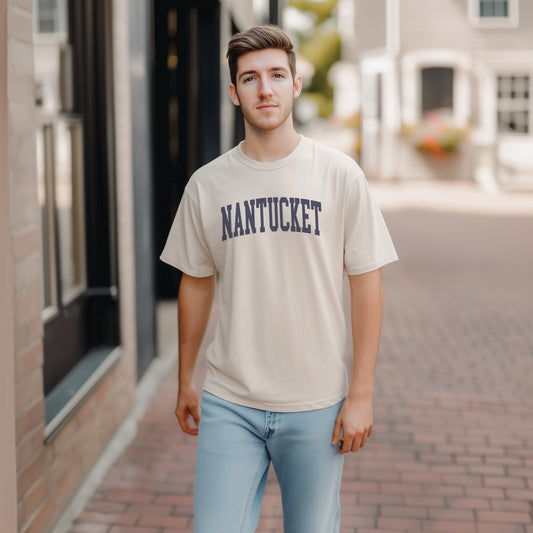 A model wears the Ivory Adult Unisex Nantucket Massachusetts Varsity Comfort Colors T-shirt.  The ﻿Nantucket Massachusetts Varsity﻿ graphic is in bold Navy in a Collegiate Block style.