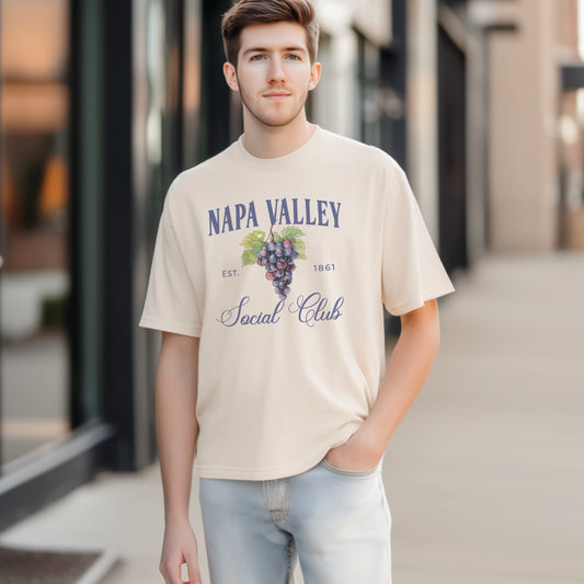 A model wears the Ivory Adult Unisex Napa Valley Wineries Social Club T-shirt.  The Napa Valley grapes  graphic is in bold purple and green in a  preppy old money style. 