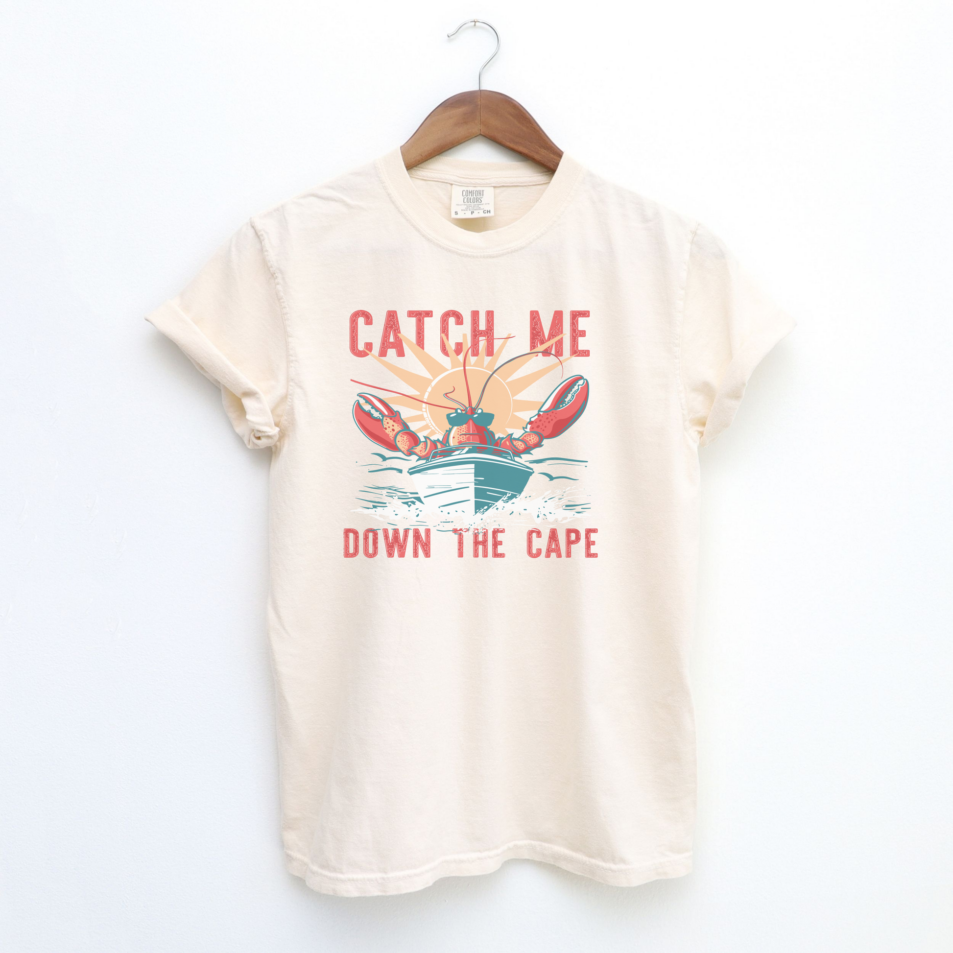 The Ivory Adult Unisex Catch Me Down The Cape Comfort Colors T-shirt lays flat on a white background. The ﻿Catch Me Down The Cape﻿ graphic is in bold multicolor in a Vintage style.