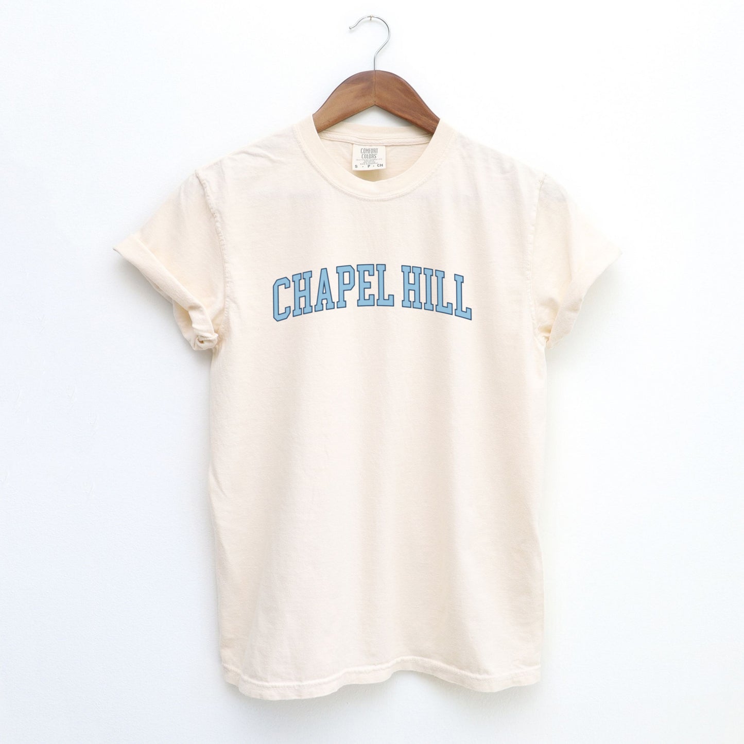 The Ivory Adult Unisex Chapel Hill Varsity Comfort Colors T-shirt lays flat on a white background. The ﻿Chapel Hill Varsity﻿ graphic is in bold Lifestyle in a Collegiate style.