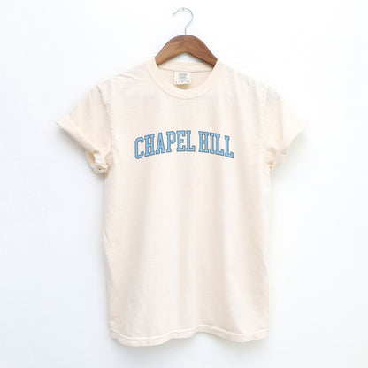The Ivory Adult Unisex Chapel Hill Varsity Comfort Colors T-shirt lays flat on a white background. The ﻿Chapel Hill Varsity﻿ graphic is in bold Lifestyle in a Collegiate style.