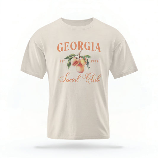 The Ivory Adult Unisex Georgia Peach Social Club T-shirt stands against a white background.  The Georgia Peach  graphic is in bold orange and green in a  preppy old money style. 