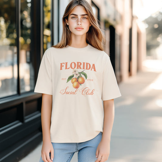A model wears the Ivory Adult Unisex Florida Social Club T-shirt.  The Florida graphic is in bold orange and green in a  preppy old money style. 