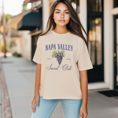 A model wears the Ivory Adult Unisex Napa Valley Wineries Social Club T-shirt.  The Napa Valley grapes  graphic is in bold purple and green in a  preppy old money style. 