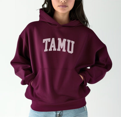 A model wears the Maroon Adult Unisex Texas A&M TAMU Hooded Sweatshirt.  The ﻿Texas A&M TAMU﻿ graphic is in bold White in a Collegiate style.