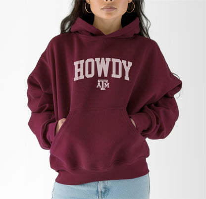 A model wears the Maroon Adult Unisex Texas A&M Howdy Varsity Hooded Sweatshirt.  The ﻿Texas A&M Howdy Varsity﻿ graphic is in bold White in a Collegiate style.