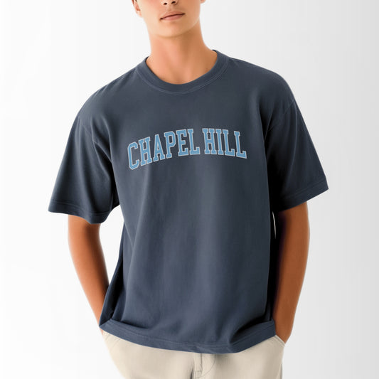 A model wears the Navy Adult Unisex Chapel Hill Varsity Comfort Colors T-shirt.  The ﻿Chapel Hill Varsity﻿ graphic is in bold Lifestyle in a Collegiate style.