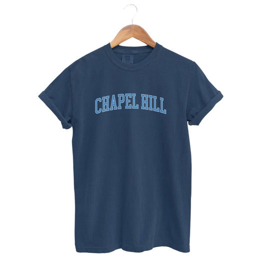 The Navy Adult Unisex Chapel Hill Varsity Comfort Colors T-shirt lays flat on a white background. The ﻿Chapel Hill Varsity﻿ graphic is in bold Lifestyle in a Collegiate style.