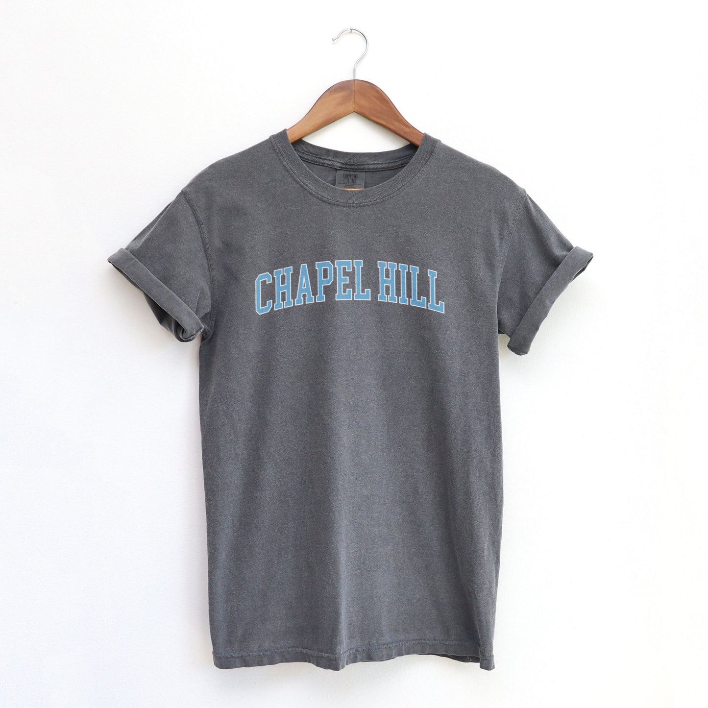 The Pepper Adult Unisex Chapel Hill Varsity Comfort Colors T-shirt lays flat on a white background. The ﻿Chapel Hill Varsity﻿ graphic is in bold Lifestyle in a Collegiate style.