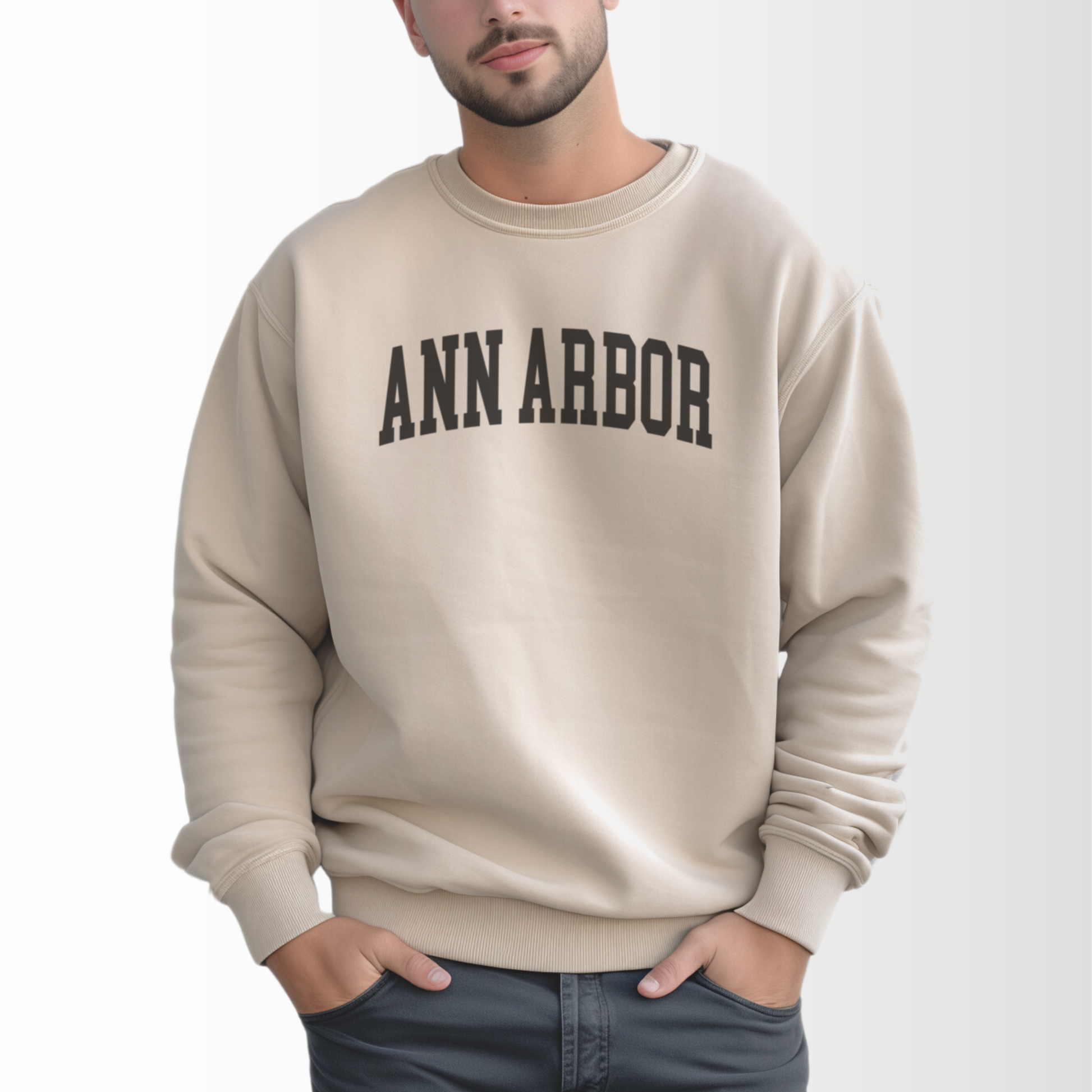 A model wears the Sand Adult Unisex Ann Arbor Michigan Varsity Crewneck Sweatshirt. The Ann Arbor Michigan Varsity graphic is in bold Black in a Collegiate style.