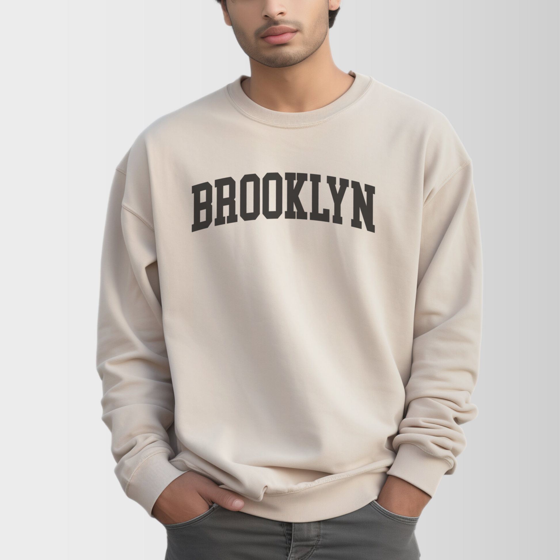 A model wears the Sand Adult Unisex Brooklyn NY Collegiate Crewneck Sweatshirt.  The ﻿Brooklyn NY Collegiate﻿ graphic is in bold Black in a Varsity style.