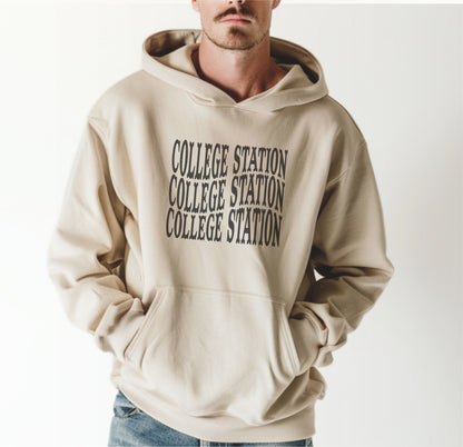 A model wears the Sand Adult Unisex College Station Western Hooded Sweatshirt.  The ﻿College Station Western﻿ graphic is in bold Black in a Western style.