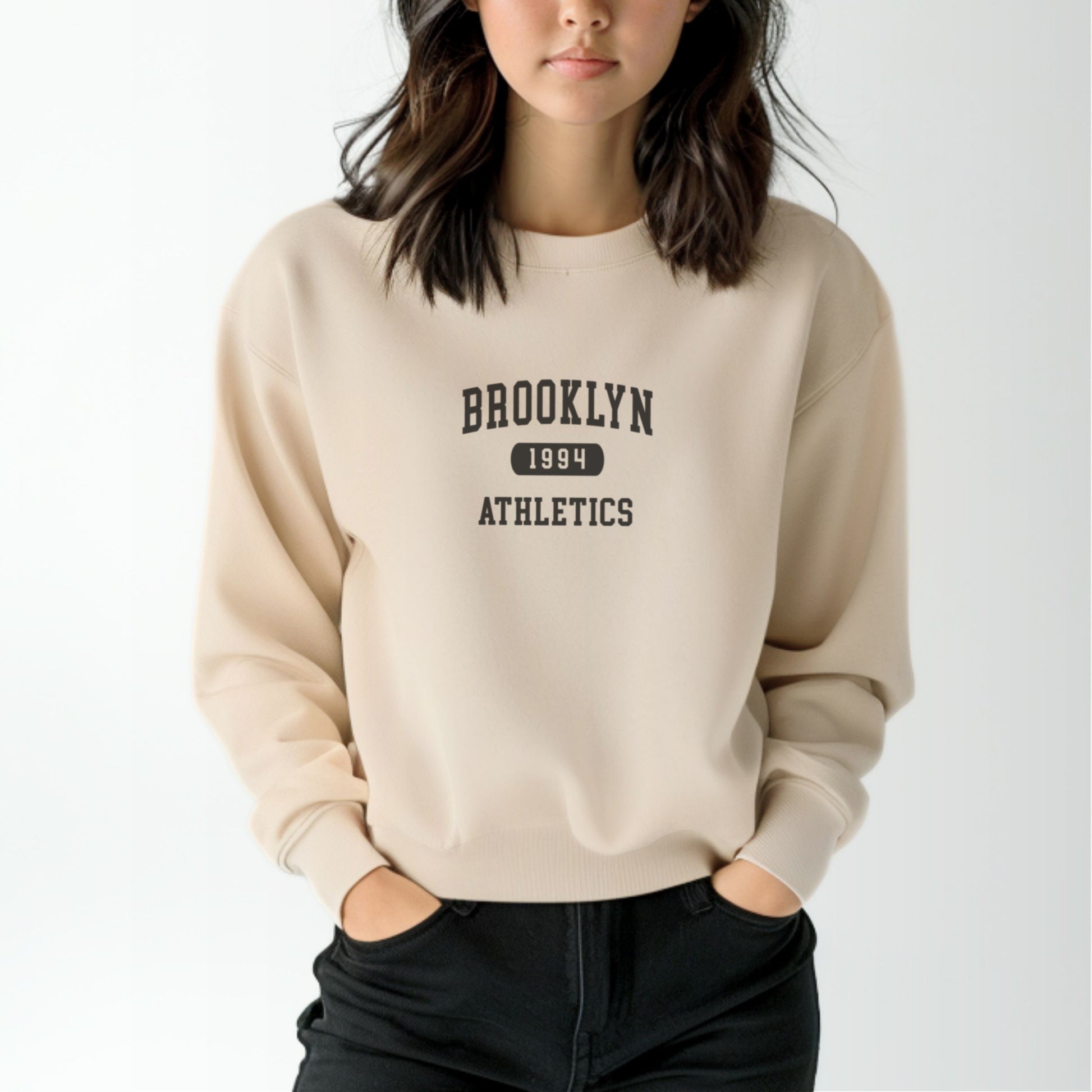 A model wears the Sand Brooklyn Athletics Crewneck Sweatshirt.  The ﻿Brooklyn Athletics﻿ graphic is in bold Navy in a Varsity style.