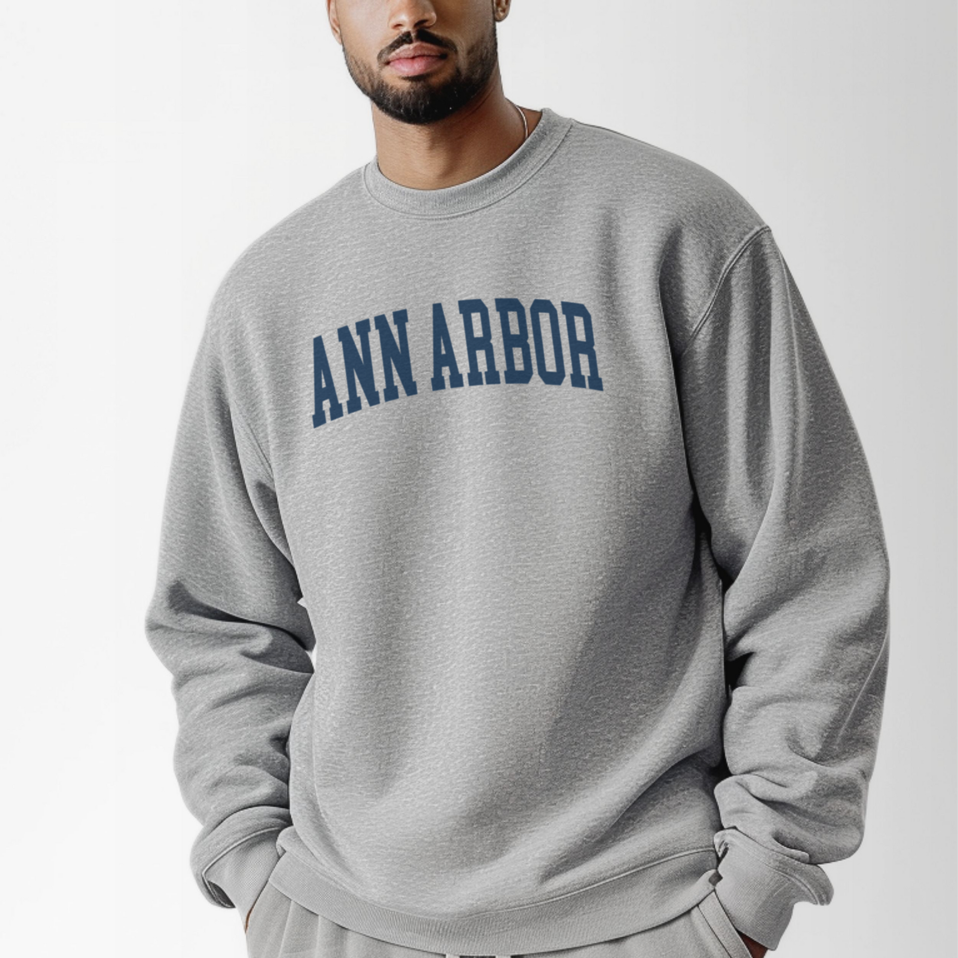 A model wears the Sport Grey Adult Unisex Ann Arbor Michigan Varsity Crewneck Sweatshirt.  The Ann Arbor Michigan Varsity graphic is in bold Navy in a Collegiate style.