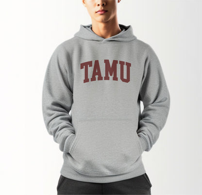 A model wears the Sport Grey Adult Unisex Texas A&M TAMU Hooded Sweatshirt.  The ﻿Texas A&M TAMU﻿ graphic is in bold Maroon in a Collegiate style.