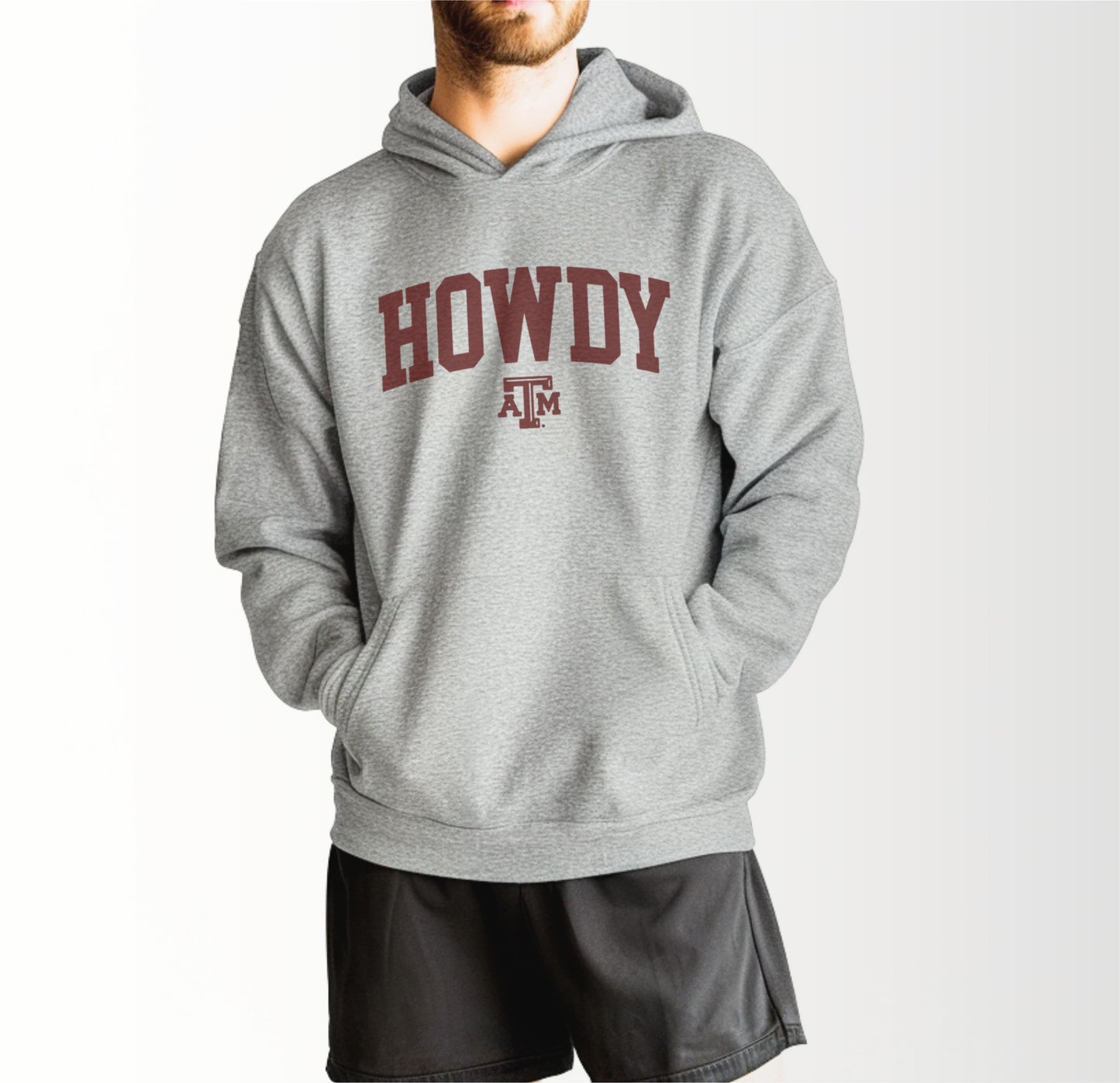 A model wears the Sport Grey Adult Unisex Texas A&M Howdy Varsity Hooded Sweatshirt.  The ﻿Texas A&M Howdy Varsity﻿ graphic is in bold Maroon in a Collegiate style.