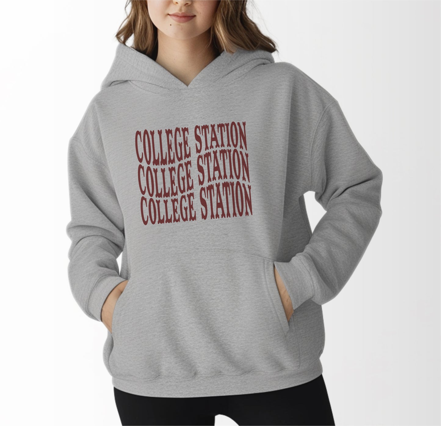 A model wears the Sport Grey Adult Unisex College Station Western Hooded Sweatshirt.  The ﻿College Station Western﻿ graphic is in bold Maroon in a Western style.