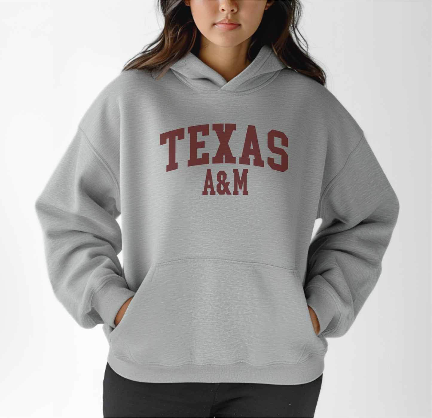 A model wears the Sport Grey Adult Unisex Texas A&M Varsity Hooded Sweatshirt.  The ﻿Texas A&M Varsity﻿ graphic is in bold Maroon in a Collegiate style.