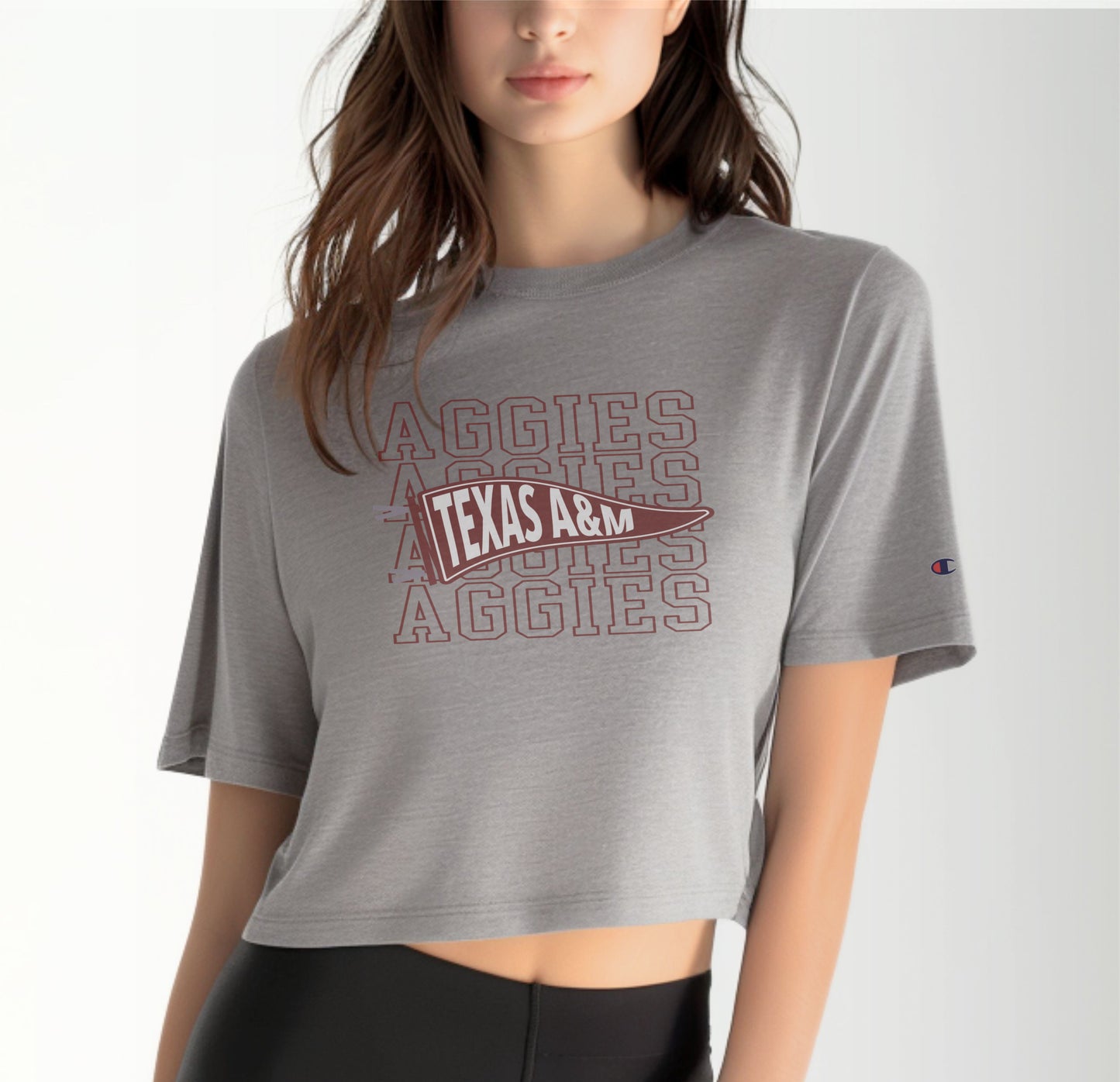 A model wears the Oxford Grey Adult Womens Texas A&M Aggies Pennant Crop Top.  The ﻿Texas A&M Aggies Pennant﻿ graphic is in bold Maroon in a Varsity style.