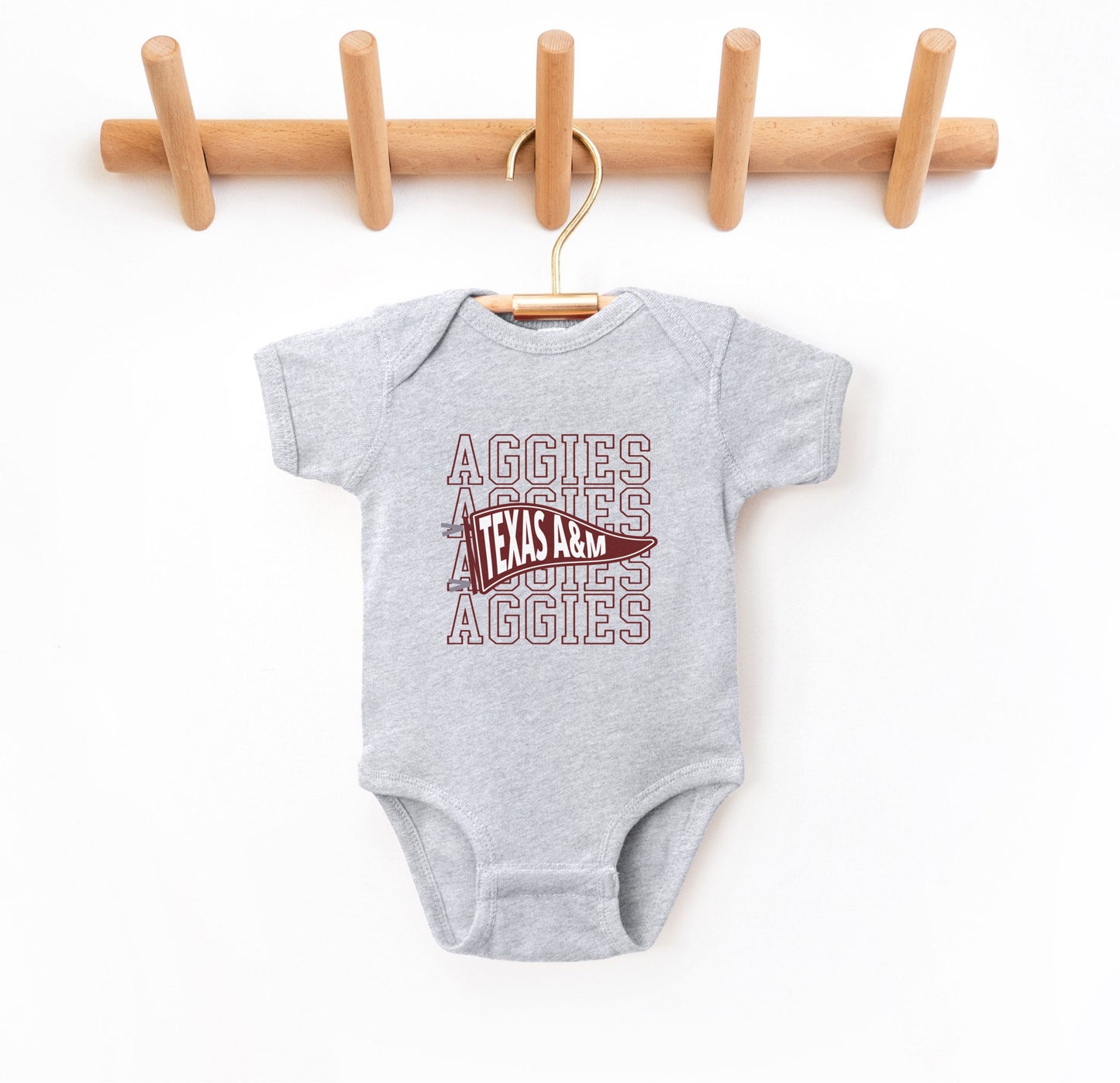 The Heather Grey Infant Unisex Texas A&M Aggies Pennant Bodysuit lays flat on a white background. The ﻿Texas A&M Aggies Pennant﻿ graphic is in bold Maroon in a Varsity style.