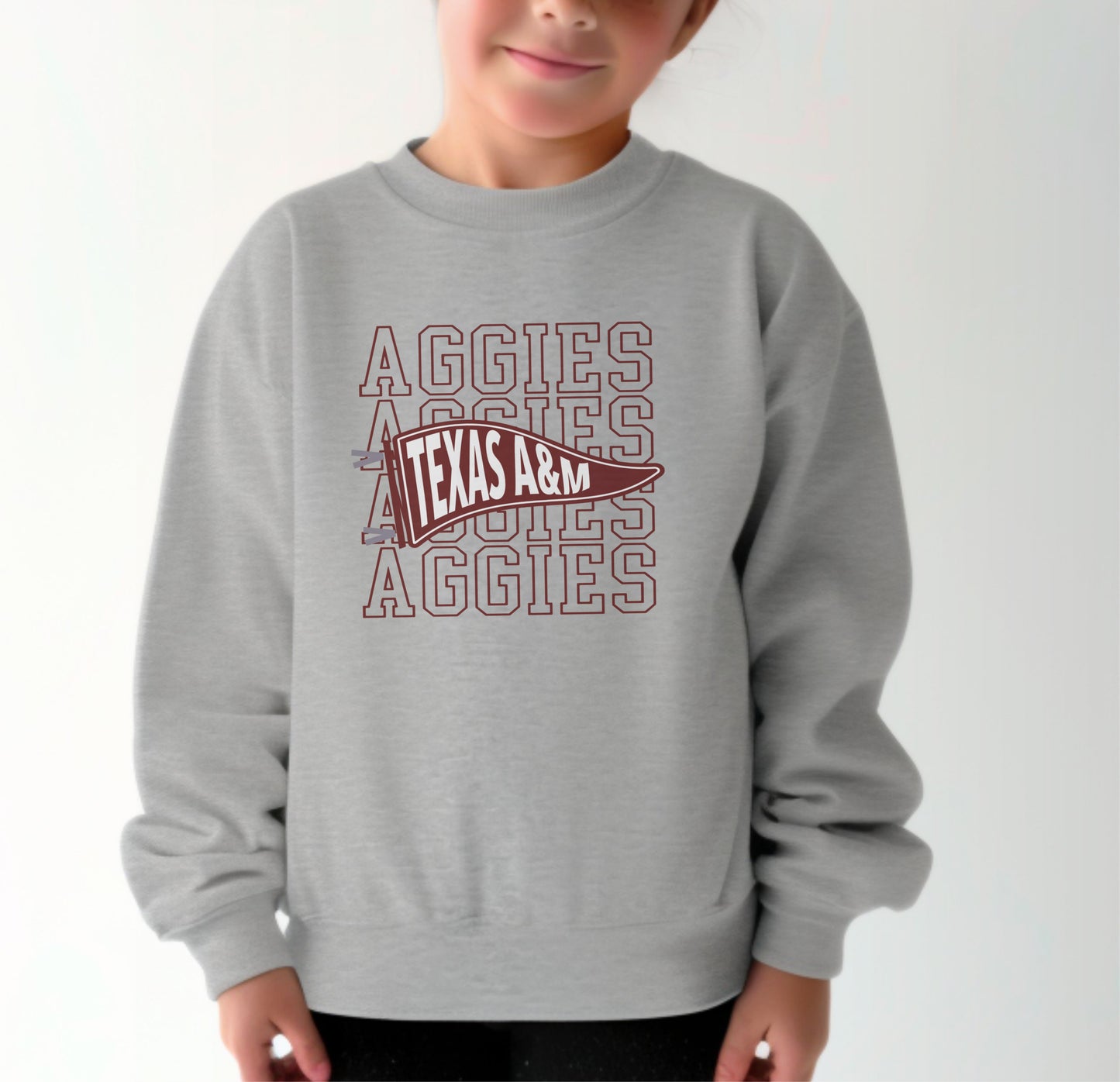 A model wears the Sport Grey Youth Unisex Texas A&M Aggies Pennant Crewneck Sweatshirt.  The ﻿Texas A&M Aggies Pennant﻿ graphic is in bold Maroon in a Varsity style.