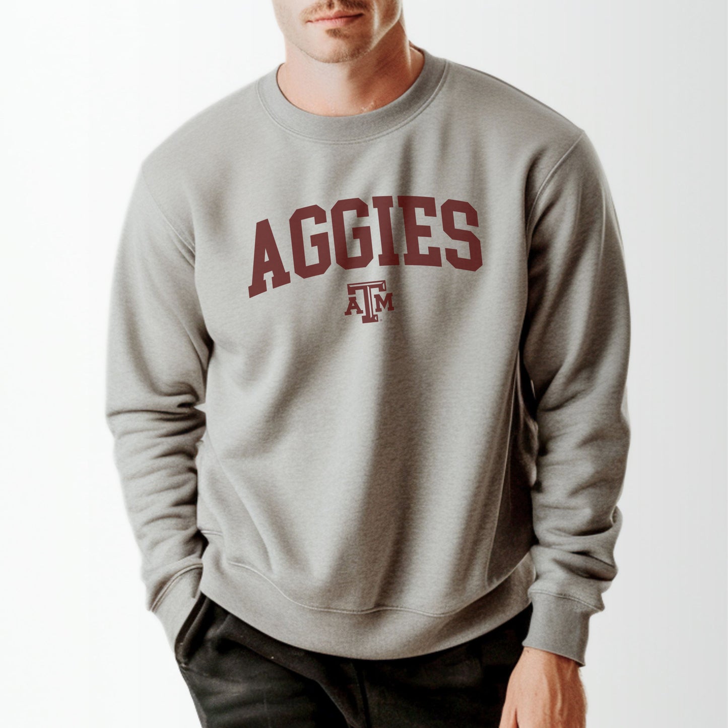 A model wears the Sport Grey Adult Unisex Texas A&M Aggies Collegiate Crewneck Sweatshirt.  The ﻿Texas A&M Aggies Collegiate﻿ graphic is in bold Maroon in a Varsity style.