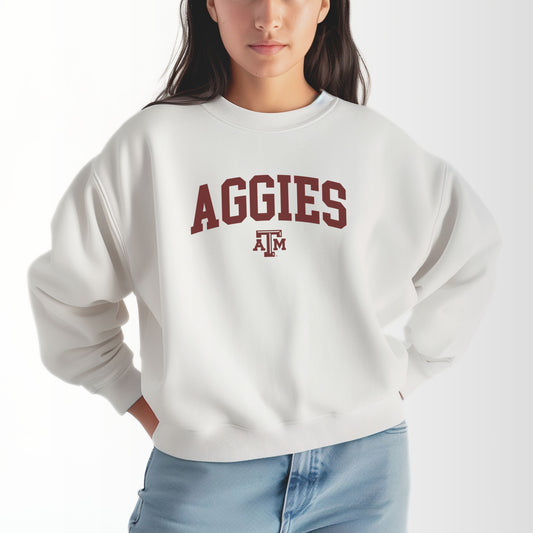 A model wears the White Adult Unisex Texas A&M Aggies Collegiate Crewneck Sweatshirt.  The ﻿Texas A&M Aggies Collegiate﻿ graphic is in bold Maroon in a Varsity style.