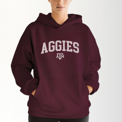 A model wears the Maroon Adult Unisex Texas A&M Aggies Collegiate Hooded Sweatshirt.  The ﻿Texas A&M Aggies Collegiate﻿ graphic is in bold White in a Varsity style.