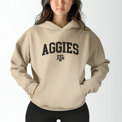 A model wears the Sand Adult Unisex Texas A&M Aggies Collegiate Hooded Sweatshirt.  The ﻿Texas A&M Aggies Collegiate﻿ graphic is in bold Black in a Varsity style.