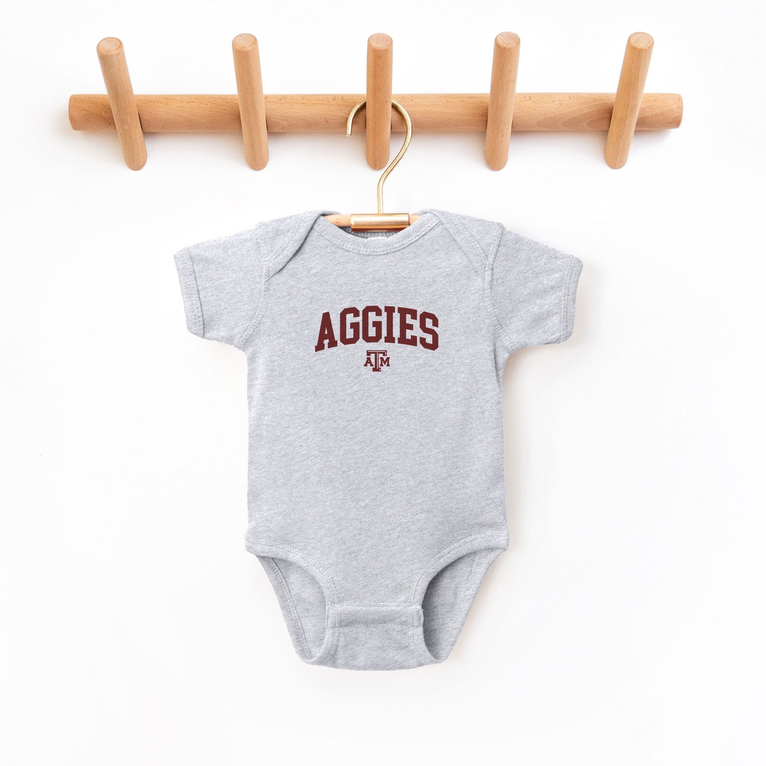 The Heather Grey Infant Unisex Texas A&M Aggies Collegiate Bodysuit lays flat on a white background. The ﻿Texas A&M Aggies Collegiate﻿ graphic is in bold Maroon in a Varsity style.