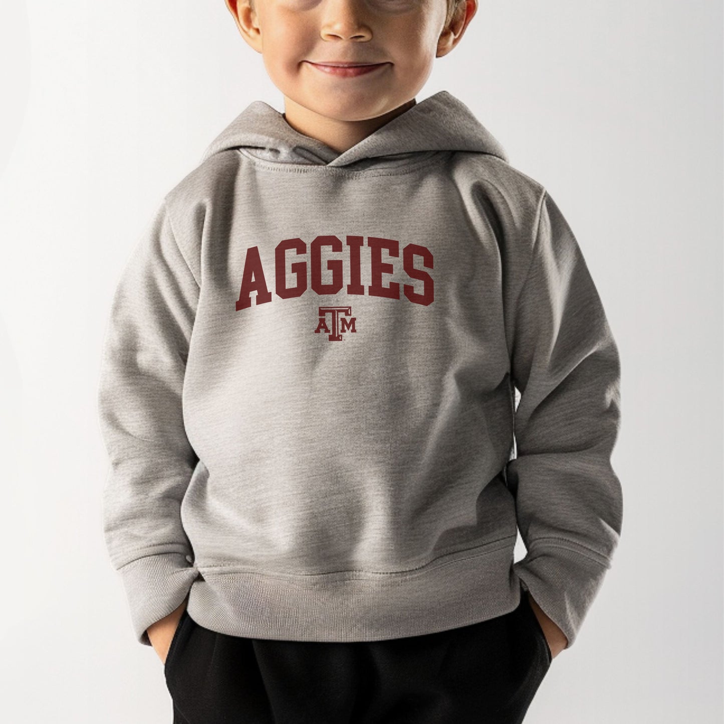 A model wears the Heather Grey Toddler Unisex Texas A&M Aggies Collegiate Hooded Sweatshirt.  The ﻿Texas A&M Aggies Collegiate﻿ graphic is in bold Maroon in a Varsity style.