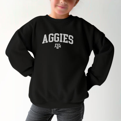 A model wears the Black Youth Unisex Texas A&M Aggies Collegiate Crewneck Sweatshirt.  The ﻿Texas A&M Aggies Collegiate﻿ graphic is in bold White in a Varsity style.