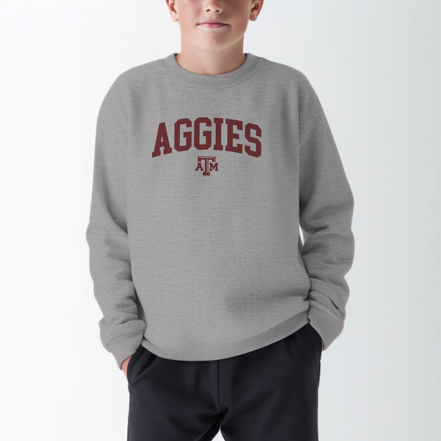 A model wears the Sport Grey Youth Unisex Texas A&M Aggies Collegiate Crewneck Sweatshirt.  The ﻿Texas A&M Aggies Collegiate﻿ graphic is in bold Maroon in a Varsity style.