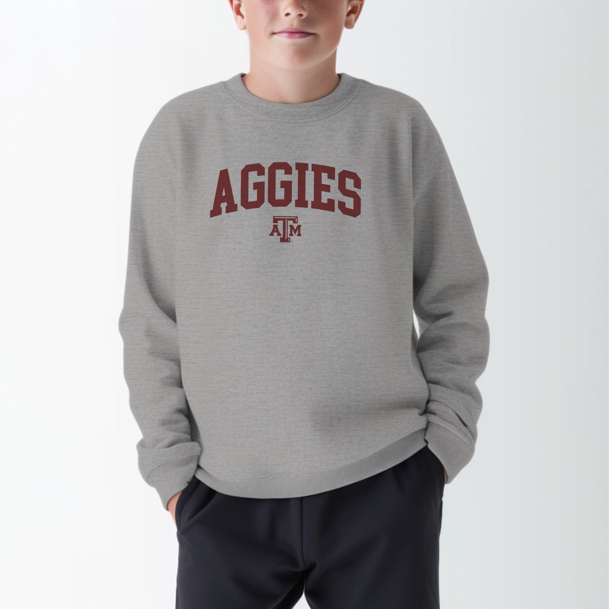 A model wears the Sport Grey Youth Unisex Texas A&M Aggies Collegiate Crewneck Sweatshirt.  The ﻿Texas A&M Aggies Collegiate﻿ graphic is in bold Maroon in a Varsity style.