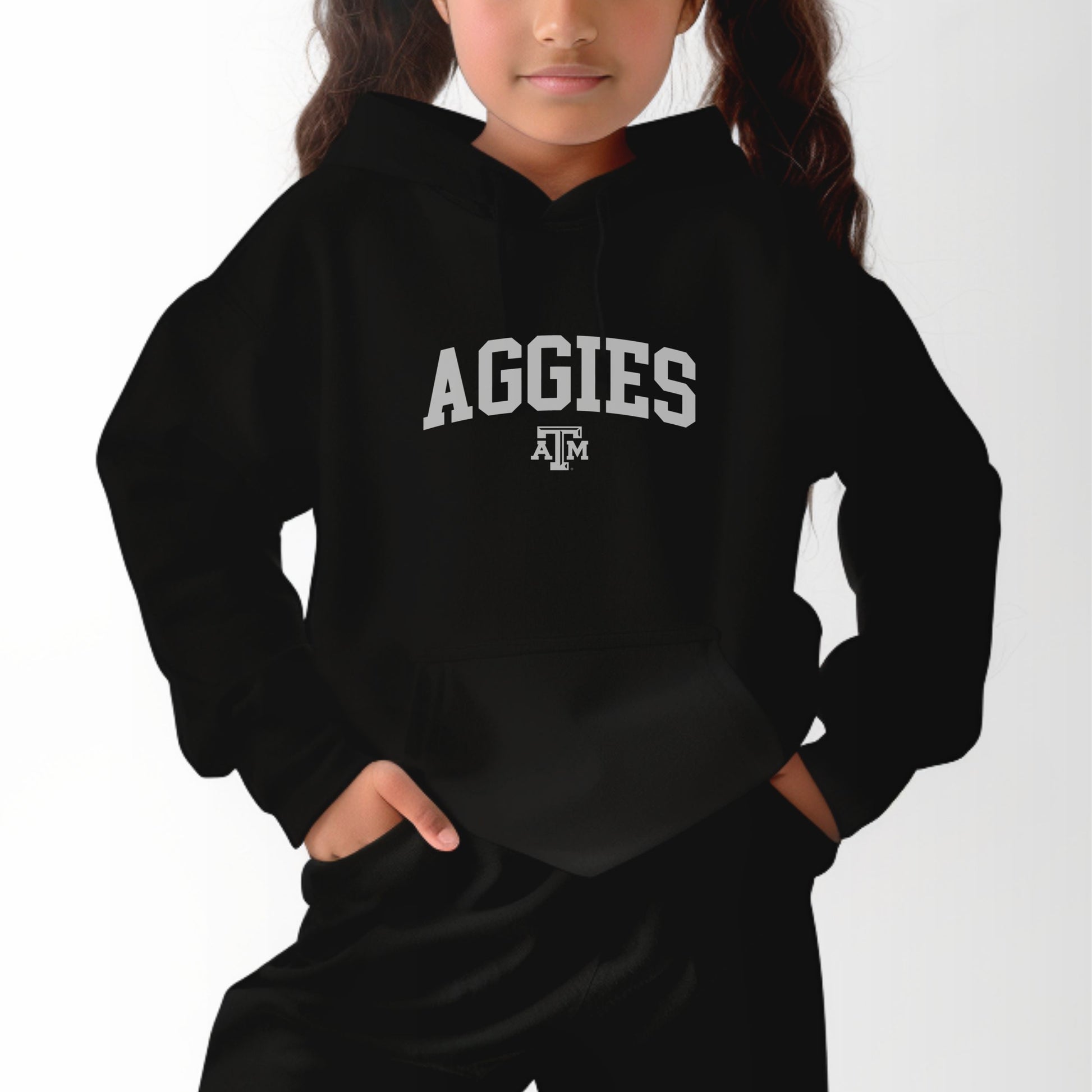 A model wears the Black Youth Unisex Texas A&M Aggies Collegiate Hooded Sweatshirt.  The ﻿Texas A&M Aggies Collegiate﻿ graphic is in bold White in a Varsity style.