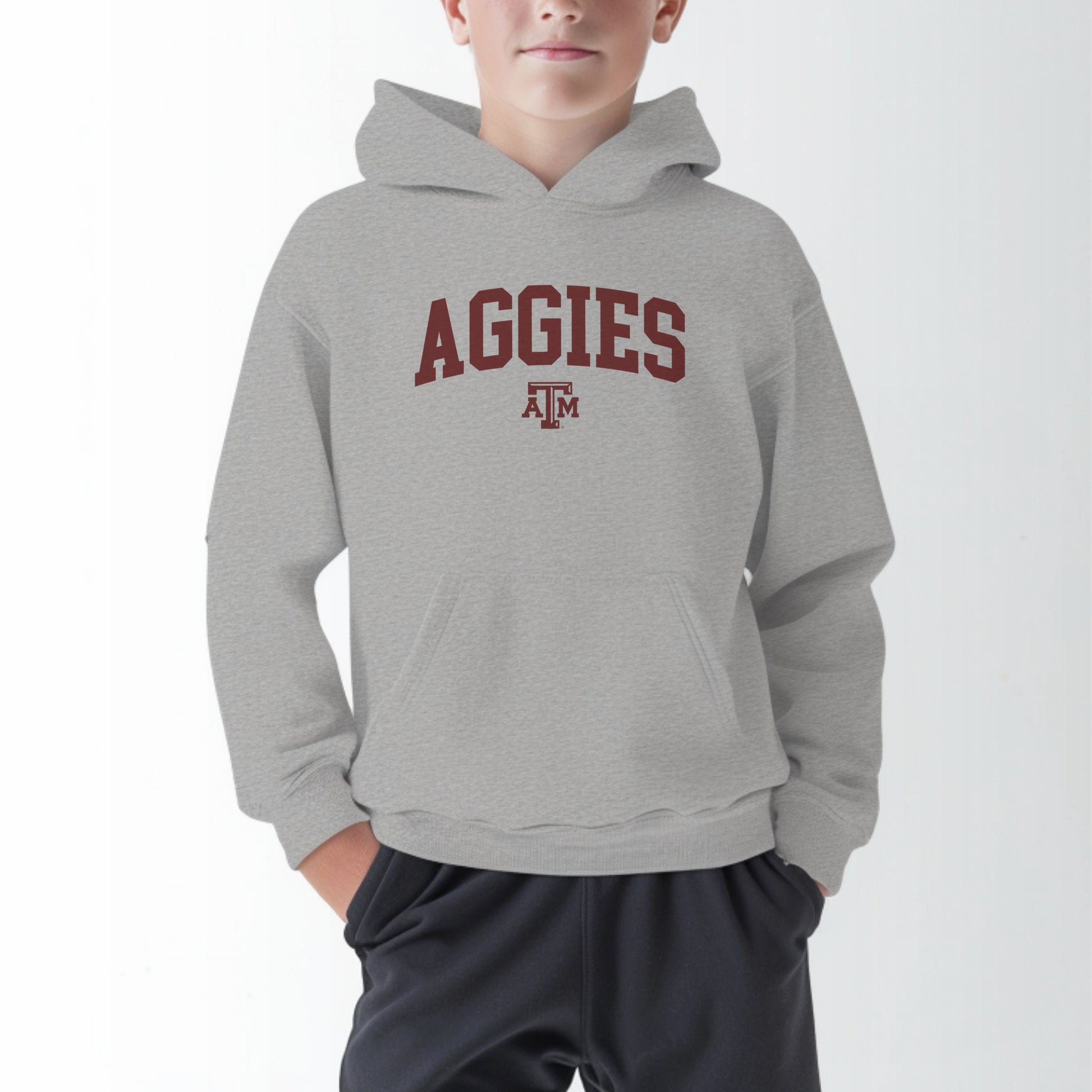 A model wears the Sport Grey Youth Unisex Texas A&M Aggies Collegiate Hooded Sweatshirt.  The ﻿Texas A&M Aggies Collegiate﻿ graphic is in bold Maroon in a Varsity style.