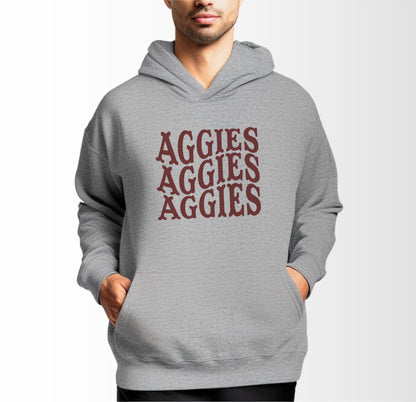 A model wears the Sport Grey Adult Unisex Texas A&M Aggies Aggies Aggies Western Hooded Sweatshirt.  The ﻿Texas A&M Aggies Aggies Aggies Western﻿ graphic is in bold Maroon in a Western style.