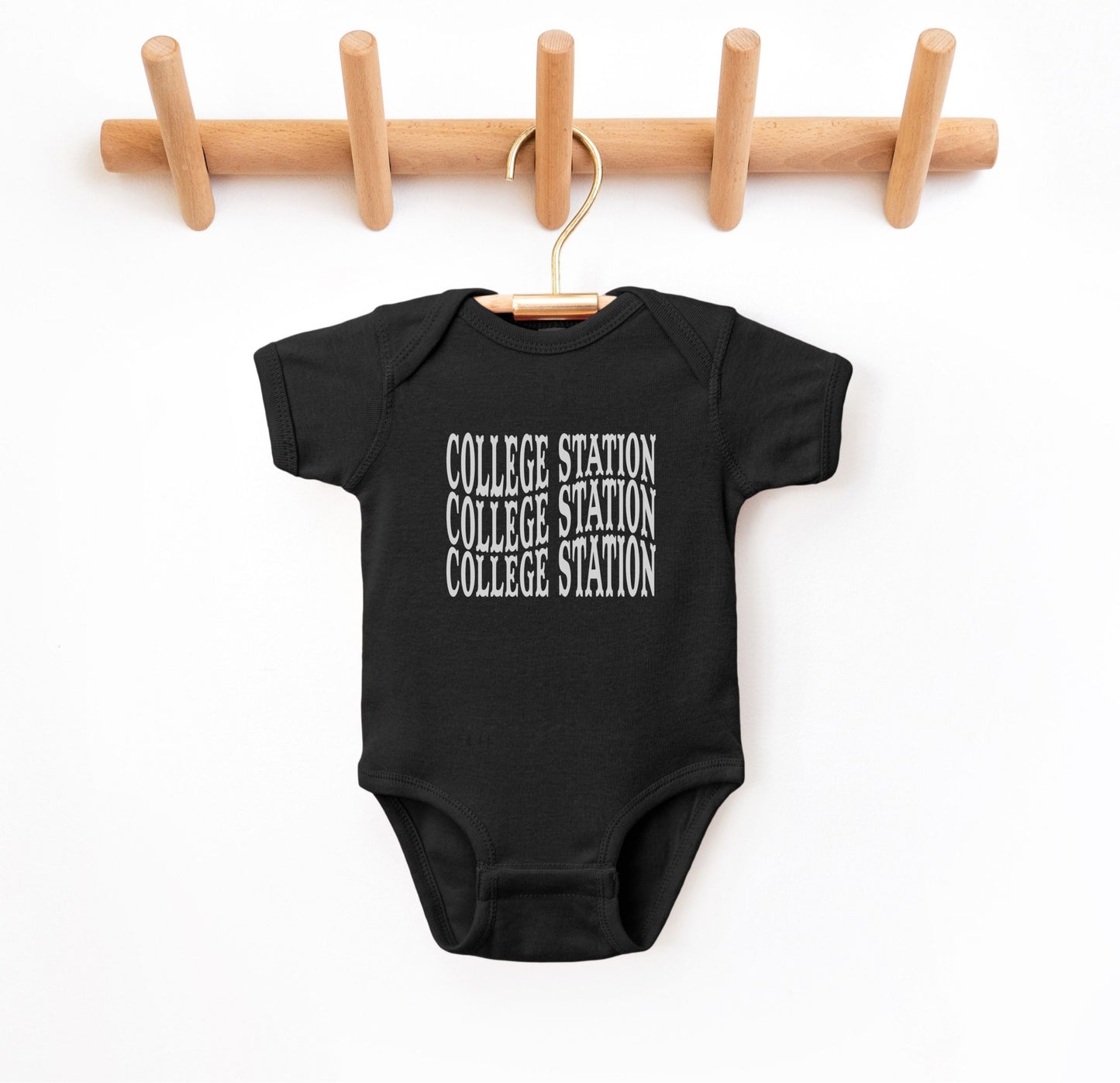 The Black Infant Unisex College Station Western Bodysuit lays flat on a white background. The ﻿College Station Western﻿ graphic is in bold White in a Western style.