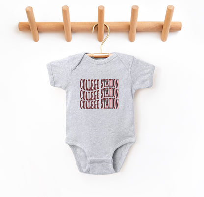 The Heather Grey Infant Unisex College Station Western Bodysuit lays flat on a white background. The ﻿College Station Western﻿ graphic is in bold Maroon in a Western style.